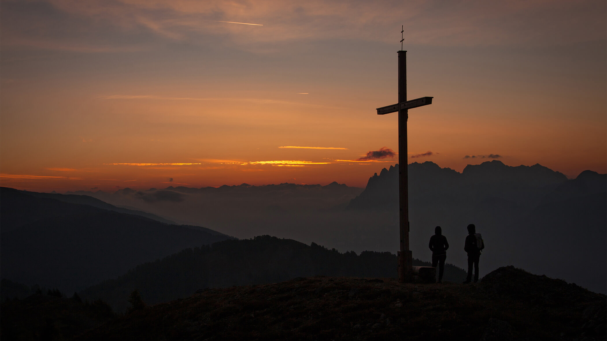 Zwei Personen stehen neben einem Gipfelkreuz bei Sonnenuntergang vor einer atemberaubender Bergkulisse - sil132 apartments