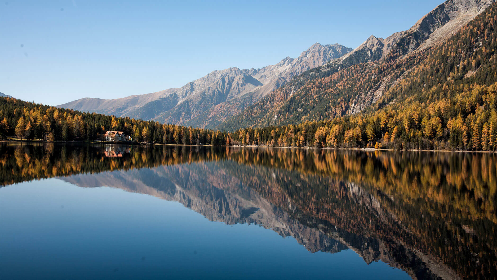 See spiegelt bunte Bäume und Berge und links ein Seehaus - sil132 apartments