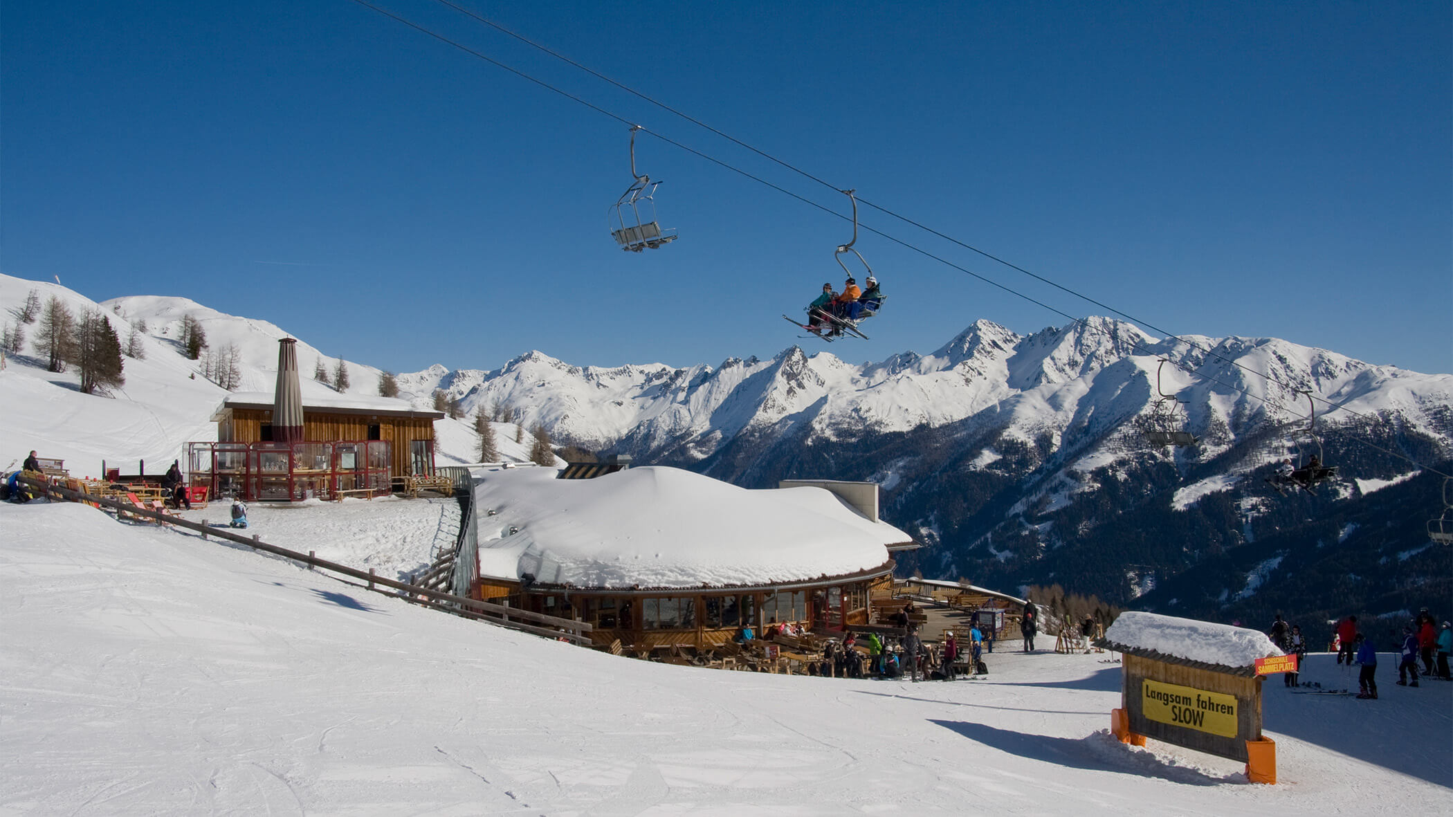 Skipiste mit Skihütte, auf dem Sessellift sitzen 3 Personen, im Hintergrund schneebedeckte Berge - sil132 apartments
